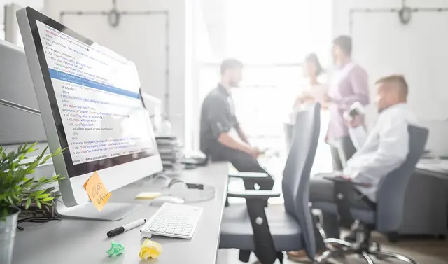 Modern office workspace with a desktop monitor displaying HTML code in the foreground, while a group of professionals discusses SEO strategy and keyword research in the blurred background.
