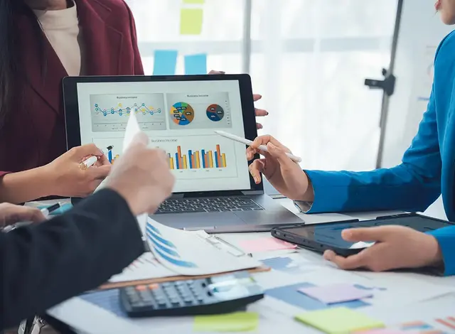 Business team reviewing SEO & PPC analytics charts on a laptop during a strategy meeting, with documents, tablet, and calculator on the table.
