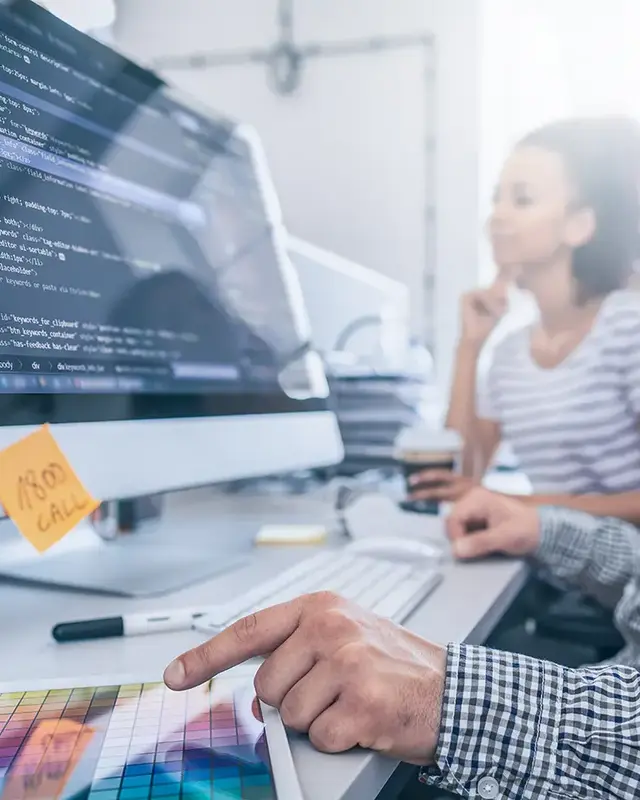Male website developer working on code at a desktop computer with a colour swatch in front of him, while a female SEO consultant sits beside him holding a coffee cup in a bright, modern office.