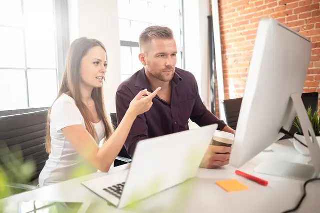 Two digital marketing professionals collaborating at a desk with laptops and a desktop monitor, discussing technical SEO strategy in a modern office setting.