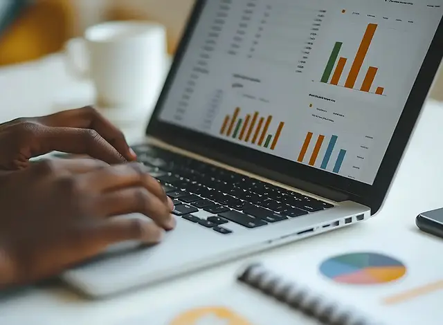 Person typing on a laptop displaying colourful bar charts and data visualisations, with a notebook and coffee cup on the desk.