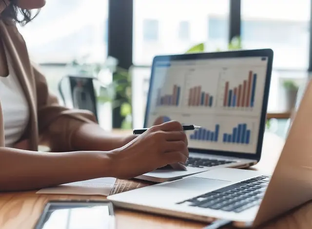 Business professional analysing bar charts and performance graphs on a laptop, using a pen to point at the screen during a data review.