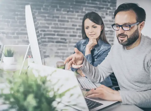 Two colleagues discussing SEO results while viewing a laptop screen in a modern office with exposed brick wall; another team member is seen thinking in the background.