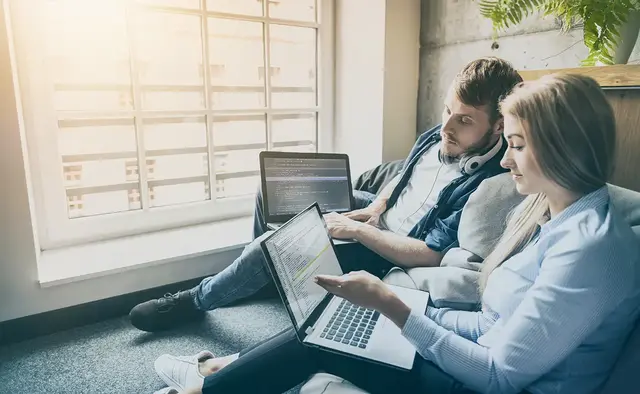 Two web designers collaborating on landing page code while working on laptops near a bright window.