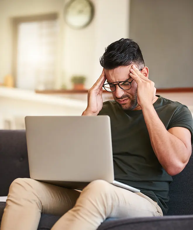 Man sitting on a sofa holding his head in frustration while looking at a laptop during a website outage.