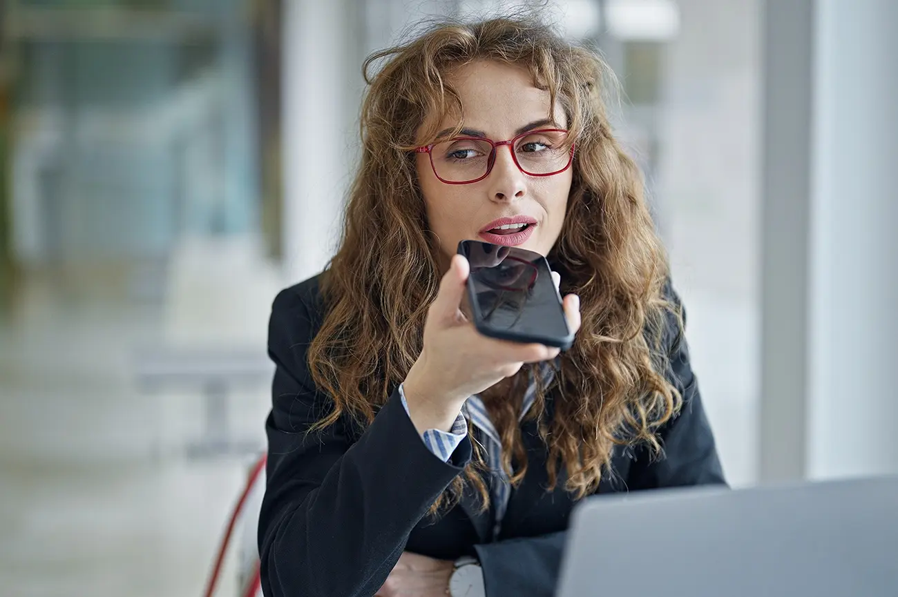 Business professional speaking into a smartphone to send a voice message while working on a laptop in a modern office – get in touch by call, email or message.