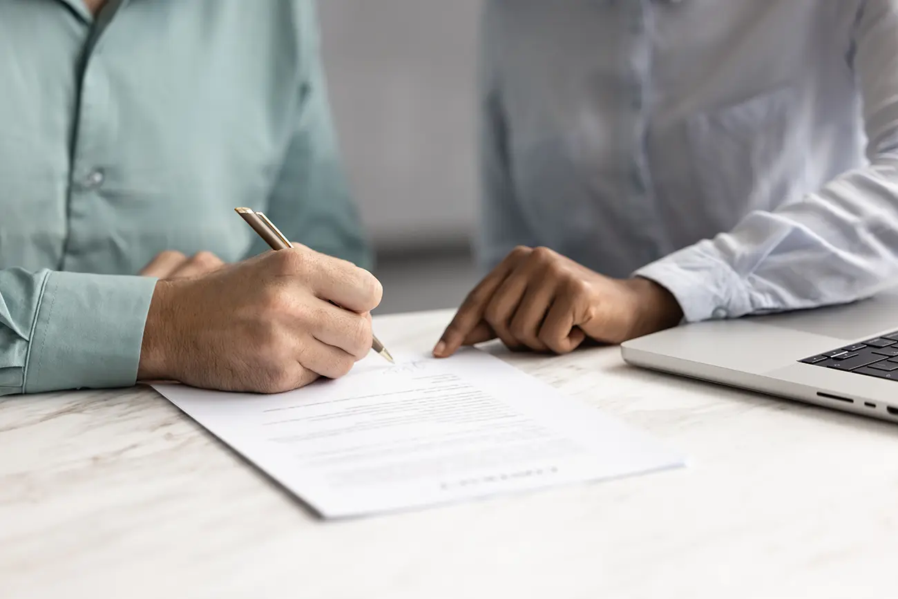 Close up of a client signing an agreement for online digital marketing services while a consultant points to the contract beside a laptop.