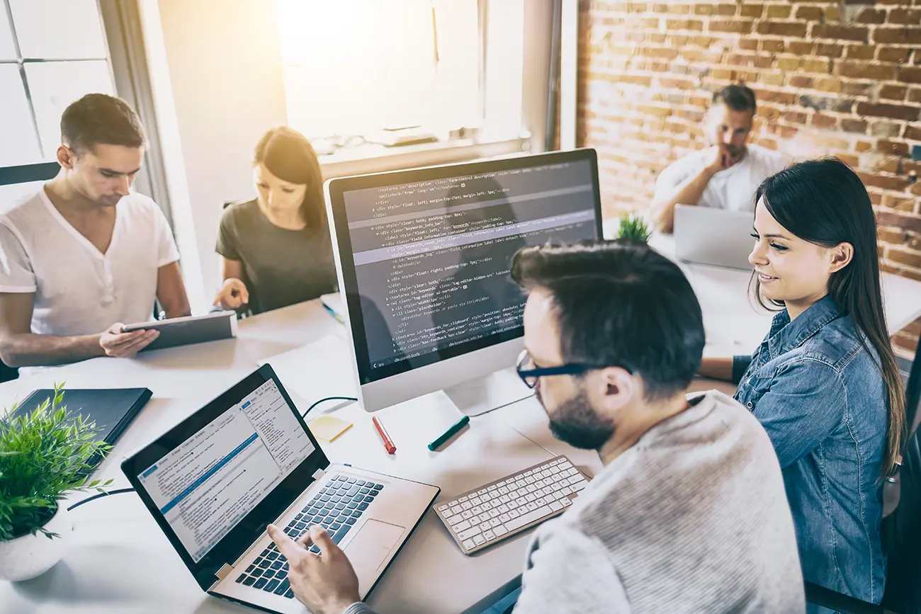 Team of digital professionals collaborating in a modern office, with laptops and monitors displaying code and analytics.